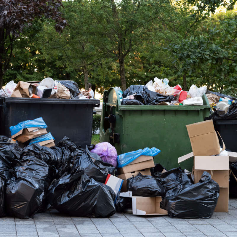 Das Bild zeigt mehrere Müllcontainer mit je 1.100 Liter Fassungsvermögen welche stark überfüllt zur Abholung bereitstehen. Davor stehen zusätzlich zahlreiche schwarze Müllsäcke.